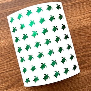 May include: A white sheet of stickers featuring a pattern of small, green sea turtles. The turtles are evenly spaced across the surface, with varying shades of green. The stickers are on a wooden surface.