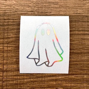 May include: A white sticker featuring a cartoon ghost with a holographic, iridescent outline. The ghost has two oval eyes and a flowing sheet-like body. The edges of the sticker have a rainbow effect.