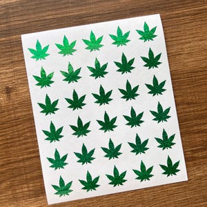 May include: A sheet of white stickers featuring a repeating pattern of green cannabis leaves. The leaves have a glittery texture and are arranged in neat rows and columns. The stickers are on a wooden surface.