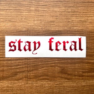 May include: A rectangular white sticker with the words "stay feral" in red gothic font. The sticker is centered on a wooden surface.