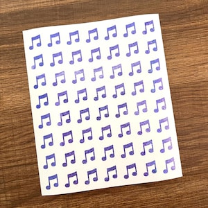 May include: A white card covered in rows of purple musical notes. The notes have a glittery texture and are evenly spaced across the card. The background is a wooden surface.