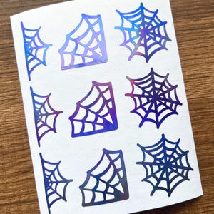 May include: A sheet of stickers featuring various spiderweb designs. The webs are in shades of blue, purple, and iridescent colors. The stickers are arranged on a white background.