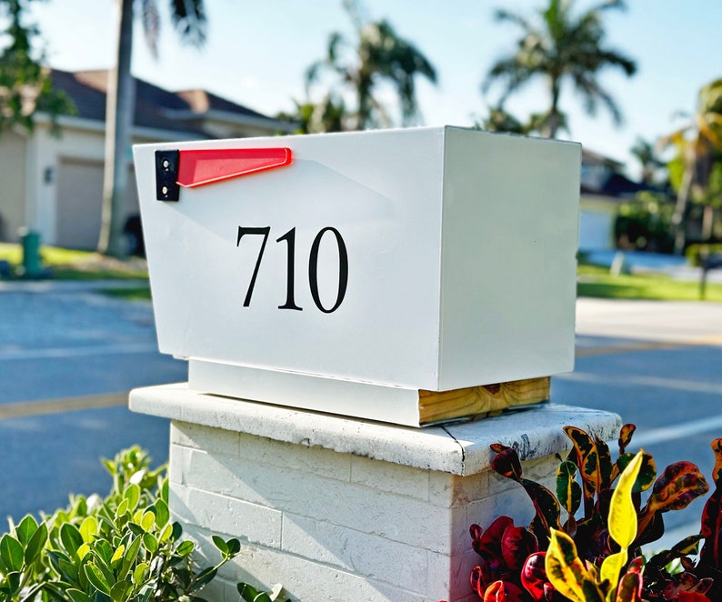 May include: White mailbox with a red flag and the number 710 on the front. The mailbox is mounted on a white brick pillar.