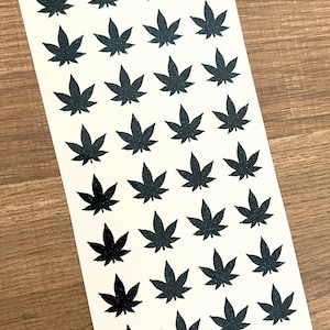 May include: A sheet of black and green glitter marijuana leaf stickers. There are 36 stickers on the sheet.