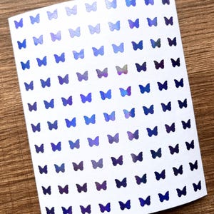 Op de afbeelding: Een vel wit papier bedekt met kleine, iriserende vlinderstickers. De vlinders zijn in tinten blauw, paars en zilver, waardoor een glinsterend effect ontstaat. De stickers zijn in nette rijen en kolommen gerangschikt.