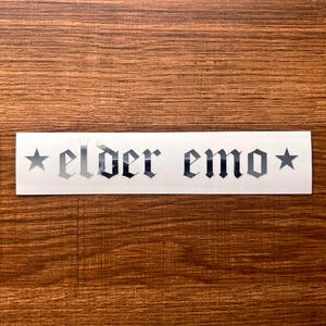 May include: A white sticker with the words "elder emo" in a gothic font, flanked by star symbols. The sticker is on a wooden surface.