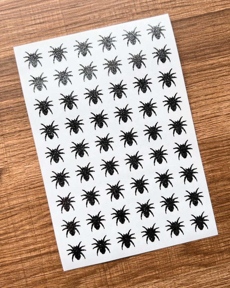 May include: A sheet of white paper covered with rows of black spider stickers. The spiders have eight legs and a round body. Some spiders have a glittery texture, while others are solid black.