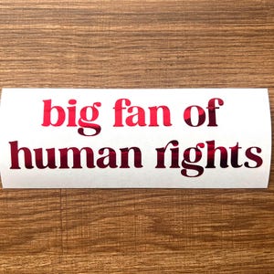 May include: A white sticker with the words "big fan of human rights" in red lettering. The sticker is rectangular with rounded corners and is placed on a wooden surface.