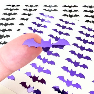 May include: A sheet of purple and black bat stickers. The stickers are shaped like bats and have a shiny, iridescent finish. A finger is holding up one of the purple bat stickers.