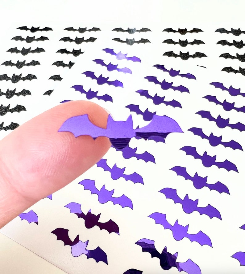 May include: A sheet of purple and black bat stickers. The stickers are shaped like bats and have a shiny, iridescent finish. A finger is holding up one of the purple bat stickers.