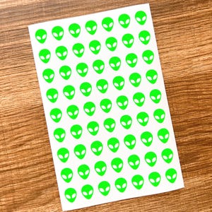 May include: A sheet of white stickers featuring a repeating pattern of bright green alien faces. The alien heads have large oval eyes and small mouths. The stickers are arranged in neat rows and columns.