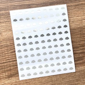 May include: A sheet of cloud-shaped stickers in various shades of gray and silver. The stickers are arranged in neat rows on a white background. The sheet is placed on a wooden surface.