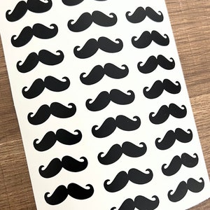 Puede incluir: Una hoja de papel blanco con 25 pegatinas de bigote negro. Las pegatinas están dispuestas en cinco filas de cinco.