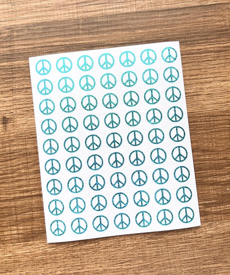 May include: A sheet of white stickers featuring a repeating pattern of teal peace symbols. The peace signs have a glittery texture, and are arranged in neat rows and columns. The stickers are on a wooden surface.
