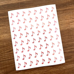 Könnte beinhalten: Eine weiße Karte mit Reihen von glitzernden roten Noten bedeckt. Die Noten sind gleichmäßig verteilt und bilden ein Muster. Die Karte liegt auf einer Holzoberfläche.