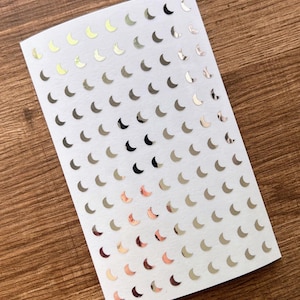 May include: A sheet of stickers featuring numerous crescent moon shapes in various metallic finishes, including gold, silver, and rose gold. The stickers are arranged on a white background.
