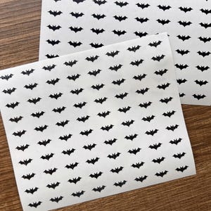 May include: Two sheets of white paper covered in small, black bat stickers. The bats are evenly spaced and have a glittery texture. The image is taken on a wooden surface, suggesting a craft or DIY project.