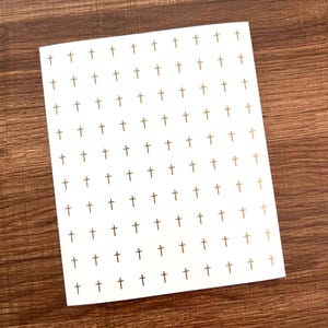 Può includere: Una carta bianca con un motivo ripetuto di croci dorate. Le croci sono uniformemente distanziate sulla superficie della carta. La carta rettangolare poggia su una superficie di legno scuro.
