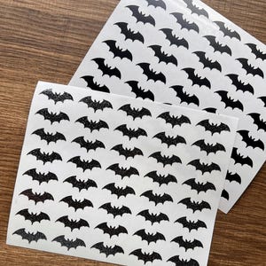 May include: Two sheets of white stickers featuring a repeating pattern of black bat silhouettes. One sheet has a glittery finish, the other is solid black. The stickers are displayed on a wooden surface.