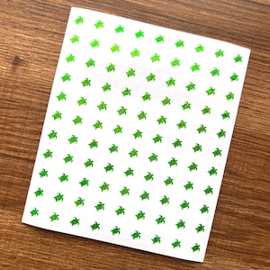 May include: A sheet of white stickers featuring a repeating pattern of small, green glittery sea turtles. The turtles are evenly spaced across the sheet, creating a playful and decorative design. The stickers are likely for crafts or embellishments.