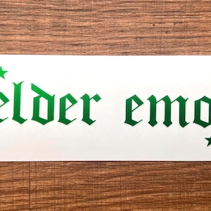 May include: A white sticker with the words "elder emo" in green gothic font, accompanied by green star accents. The sticker is rectangular with rounded corners, set against a wood grain background.