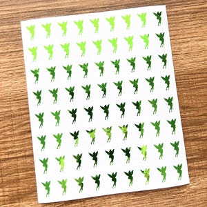 Puede incluir: Una hoja de pegatinas con un patrón repetido de pequeñas siluetas de hadas. Las hadas están representadas en varios tonos de verde, con alas extendidas, y están dispuestas sobre un fondo blanco.