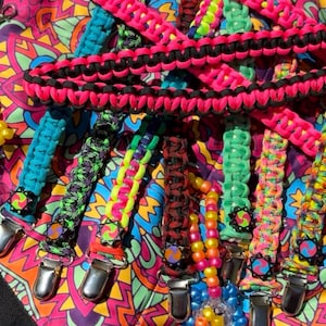 May include: A collection of colorful pacifier clips in various shades of pink, green, blue, and black. The clips are made of woven cord and feature metal clasps. The background is a vibrant, patterned fabric.
