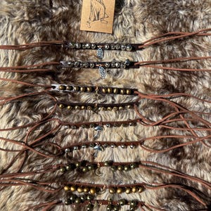 Puede incluir: Una colección de pulseras de cuero marrón con varios diseños de cuentas y abalorios metálicos. Algunas pulseras presentan abalorios plateados como hojas, lunas y estrellas. Una pequeña tarjeta con un dibujo de una persona está en el fondo.