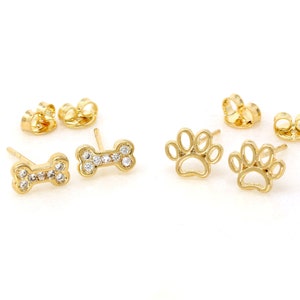May include: A set of gold-tone earrings featuring a paw print and a bone design. The bone earrings are adorned with clear cubic zirconia stones.