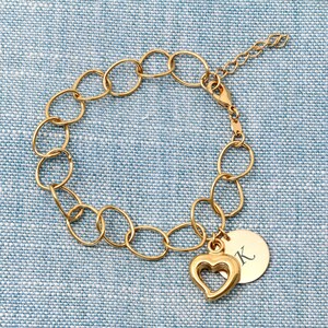 Könnte beinhalten: Ein goldenes Kettenarmband mit einem Herz-Anhänger und einem runden Anhänger, auf dem der Buchstabe "K" eingraviert ist.