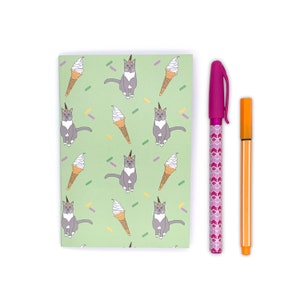 May include: A notebook with a light green background featuring a pattern of grey cats with unicorn horns and ice cream cones. A pink pen with a chevron pattern and an orange pen are next to the notebook.
