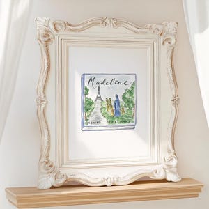 May include: A framed print of a book cover titled "Madeline" with a watercolor illustration of the Eiffel Tower and a figure in a blue coat. The frame is ornate, white, and sits on a light-colored wooden shelf.