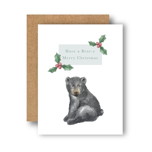 Puede incluir: Una tarjeta blanca con una ilustración de un oso negro y el texto "Have a Bear-y Merry Christmas" en un cuadro azul claro. La tarjeta está decorada con hojas de acebo verdes y bayas rojas.