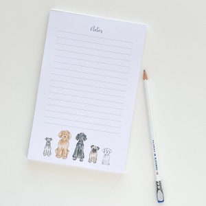 May include: A white notepad with lined pages and the word "Notes" in script at the top. The bottom of the notepad features watercolor illustrations of six different dog breeds. A white pencil with black text and a blue eraser sits to the right.