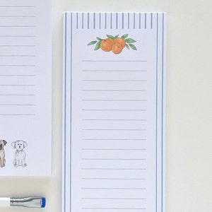 May include: A white notepad with blue vertical stripes and a watercolour illustration of oranges and leaves. The notepad has lined pages for writing notes. Another notepad with dog illustrations is partially visible.