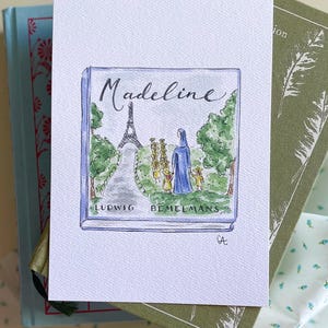 May include: A watercolor illustration of the book "Madeline" by Ludwig Bemelmans. The cover art depicts the Eiffel Tower, a nun, and several girls. The artwork is on textured paper, resting on top of two books.