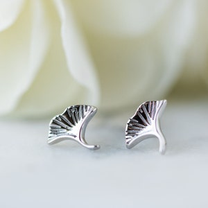 May include: A pair of silver-colored stud earrings shaped like ginkgo leaves. The earrings have detailed veining and a curved post. The background features a blurred white flower, creating a soft, elegant aesthetic.