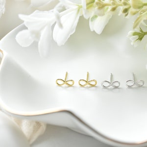 Op de afbeelding: Drie paar oorknopjes met oneindigheidssymbool in goud- en zilverkleuren worden gepresenteerd op een witte, bloemvormige schaal. De oorbellen zijn klein en delicaat, met een eenvoudig, minimalistisch ontwerp. Witte bloemen en groen zijn op de achtergrond.