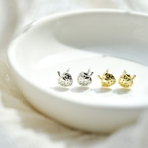 May include: Four small strawberry-shaped earrings, two silver and two gold, displayed on a white ceramic dish.