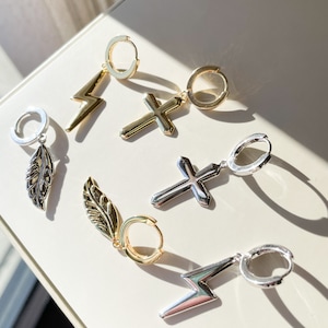 May include: Six pairs of dangle earrings with different charms. The charms include a feather, a lightning bolt, and a cross. The earrings are made of silver and gold metal.