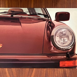 May include: Close-up of a maroon-colored vintage car, showcasing the front end. The image highlights the headlight, side mirror, and hood with the Porsche emblem. The car's design is classic and sleek, with a focus on its iconic features.