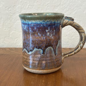 Peut inclure: Mug en céramique fait à la main avec un glaçage unique. La tasse présente une anse texturée et un dégradé de couleurs, passant d'un bord vert foncé à des tons bleus et bruns. Le glaçage crée un effet dégoulinant à l'extérieur.