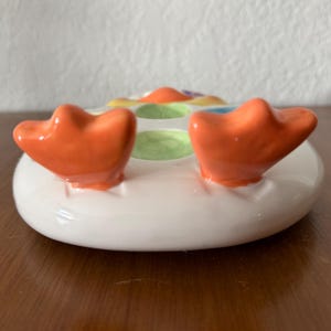 K&ouml;nnte beinhalten: Eine keramische Seifenschale in Entenform. Die Schale ist wei&szlig; mit orangefarbenen F&uuml;&szlig;en und bunten Akzenten. Die F&uuml;&szlig;e sind erh&ouml;ht und gebogen, was der Schale ein verspieltes Aussehen verleiht. Die Schale ist f&uuml;r ein St&uuml;ck Seife konzipiert.
