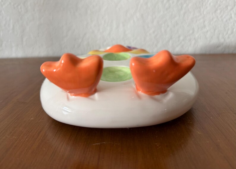 K&ouml;nnte beinhalten: Eine keramische Seifenschale in Entenform. Die Schale ist wei&szlig; mit orangefarbenen F&uuml;&szlig;en und bunten Akzenten. Die F&uuml;&szlig;e sind erh&ouml;ht und gebogen, was der Schale ein verspieltes Aussehen verleiht. Die Schale ist f&uuml;r ein St&uuml;ck Seife konzipiert.