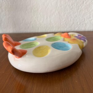 K&ouml;nnte beinhalten: Ein wei&szlig;er Eierhalter aus Keramik mit bunten Tupfen und einem K&uuml;ken-Design. Der Eierhalter hat orangefarbene F&uuml;&szlig;e und fasst mehrere Eier. Das K&uuml;ken hat einen orangefarbenen Schnabel und eine lila Blume auf dem Kopf. Der Eierhalter steht auf einer Holzoberfl&auml;che.