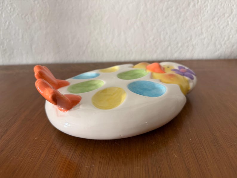 K&ouml;nnte beinhalten: Ein wei&szlig;er Eierhalter aus Keramik mit bunten Tupfen und einem K&uuml;ken-Design. Der Eierhalter hat orangefarbene F&uuml;&szlig;e und fasst mehrere Eier. Das K&uuml;ken hat einen orangefarbenen Schnabel und eine lila Blume auf dem Kopf. Der Eierhalter steht auf einer Holzoberfl&auml;che.