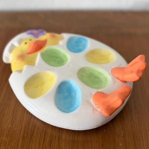 K&ouml;nnte beinhalten: Eine wei&szlig;e, keramikartige, eif&ouml;rmige Servierschale mit bunten, eif&ouml;rmigen F&auml;chern. Die Schale zeigt ein gelbes K&uuml;ken, das aus dem Ei schl&uuml;pft, und orangefarbene herzf&ouml;rmige Griffe. Die F&auml;cher sind in Gelb, Gr&uuml;n und Blau bemalt.