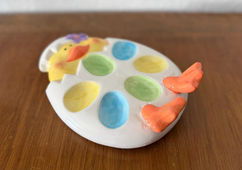 K&ouml;nnte beinhalten: Eine wei&szlig;e, keramikartige, eif&ouml;rmige Servierschale mit bunten, eif&ouml;rmigen F&auml;chern. Die Schale zeigt ein gelbes K&uuml;ken, das aus dem Ei schl&uuml;pft, und orangefarbene herzf&ouml;rmige Griffe. Die F&auml;cher sind in Gelb, Gr&uuml;n und Blau bemalt.