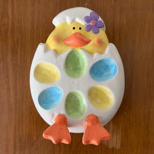 May include: A ceramic Easter egg holder in the shape of a chick hatching from an egg. The chick is yellow with an orange beak and a purple flower. The egg has six colourful egg-shaped indentations in yellow, green, and blue.