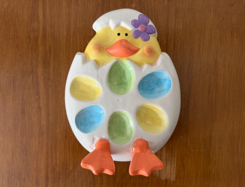 K&ouml;nnte beinhalten: Ein Keramik-Ostereierhalter in Form eines K&uuml;kens, das aus einem Ei schl&uuml;pft. Das K&uuml;ken ist gelb mit einem orangefarbenen Schnabel und einer lila Blume. Das Ei hat sechs farbige, eif&ouml;rmige Vertiefungen in Gelb, Gr&uuml;n und Blau.